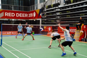 "Four badminton players compete in a doubles match on a green court at the Red Deer 2019 event, with the score 19-13 displayed."