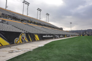 "An empty sports stadium features a green turf field, rows of gray seats, and black banners with a tiger mascot and Tim Hortons branding under a cloudy sky."