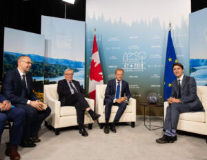 "Justin Trudeau, Donald Tusk, Jean-Claude Juncker, and Peter Praet are seated at the G7 2018 Charlevoix summit, flanked by Canadian and EU flags."