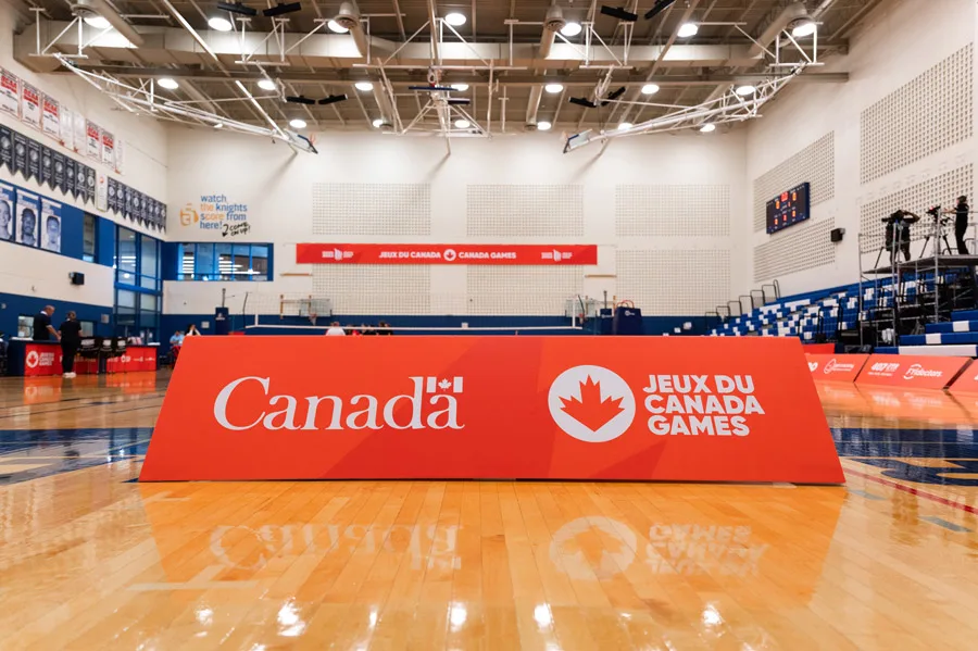 An indoor sports gymnasium features a large orange sign on the polished wooden court, displaying ""Canada"" and ""JEUX DU CANADA GAMES,"" with blue bleachers and event equipment in the background.
