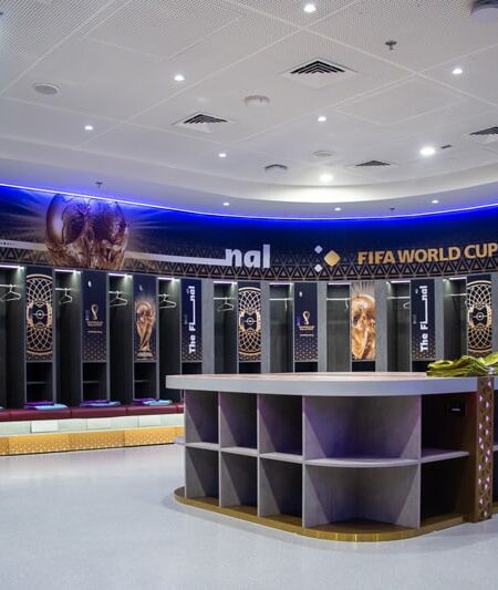 "A modern locker room lavishly decorated with FIFA World Cup Qatar 2022 branding, featuring individual stalls, maroon benches, and a large central table."