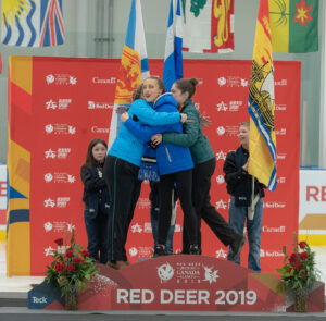 "Three young women hug on a podium at the Red Deer 2019 Canada Games, surrounded by event flags and branding."