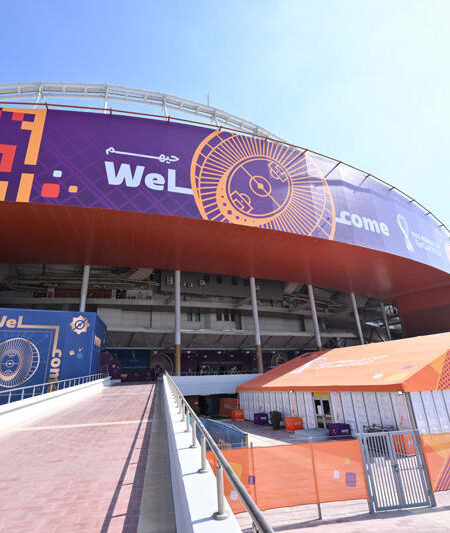"A large stadium entrance is decorated with orange and purple ""Welcome"" banners for the FIFA World Cup, with a walkway leading towards it."