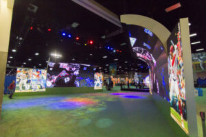An immersive baseball exhibit featuring large curved LED screens displaying players and a green floor with colorful light projections.