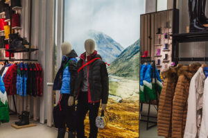 "A retail store display features mannequins in outdoor jackets, racks of colorful clothing, and boots, with a large mountain landscape mural in the background."