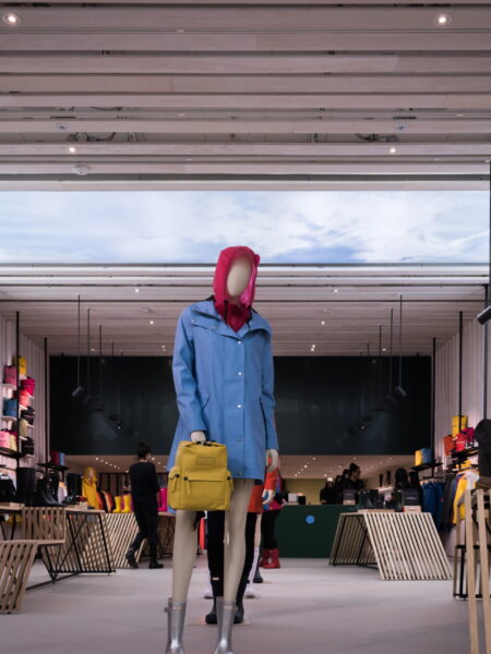 A mannequin wearing a blue raincoat and yellow backpack stands in a modern outdoor apparel store featuring other clothing displays and large scenic backdrop screens.