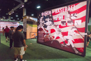"An indoor baseball exhibit features large displays about ""Women in Baseball"" and ""Negro Leagues Veterans,"" with visitors viewing them."