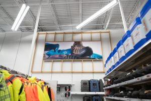 "An interior view of a retail store featuring a large advertisement for a hiking boot, surrounded by racks of work jackets and shelves of footwear and blue storage containers."