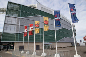 "Flags for the NHL Heritage Classic 2019, featuring the Winnipeg Jets, Calgary Flames, and Tim Hortons, are displayed outside a modern stadium."