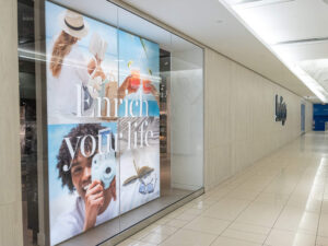 "A large glass storefront display reads ""Enrich your life"" over four panels depicting leisure activities like reading, photography, and enjoying drinks, set in a bright shopping mall hallway."
