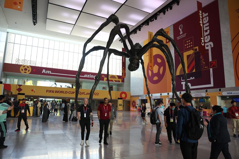 "A giant black spider sculpture stands prominently in a spacious hall decorated with FIFA World Cup Qatar 2022 branding, surrounded by people."