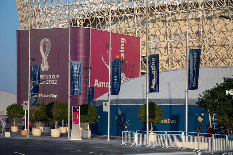 Exterior of a building wrapped with World Cup Qatar 2022 banners, blue flags, and potted trees.
