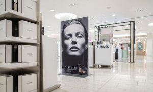 "A bright department store aisle showcases Chanel beauty product displays, including a large black and white ad for LE LIFT PRO featuring a model, and shelves filled with Chanel boxes."