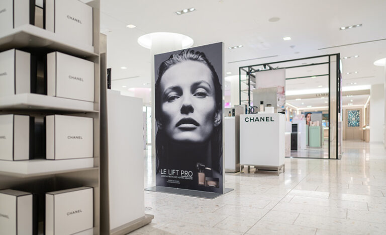 "A bright department store aisle showcases Chanel beauty product displays, including a large black and white ad for LE LIFT PRO featuring a model, and shelves filled with Chanel boxes."