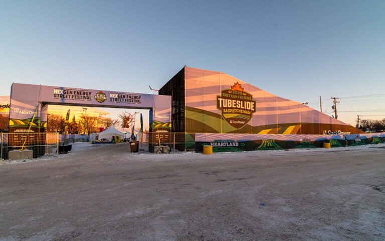 "A snowy outdoor festival features an entrance arch for the ""NexGen Energy Street Festival"" and a large structure with a ""2023 Grey Cup Festival Tubeslide Saskatchewan"" banner."