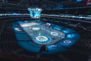 "An empty Bell MTS Place ice hockey arena is bathed in blue light, featuring the Winnipeg Jets logo on the ice and a large central scoreboard."