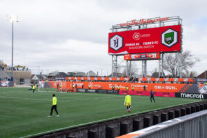 "A wide view of Tim Hortons Field shows soccer players on the pitch and a large scoreboard displaying ""Forge FC vs York9 FC Inaugural Match"" under an overcast sky."
