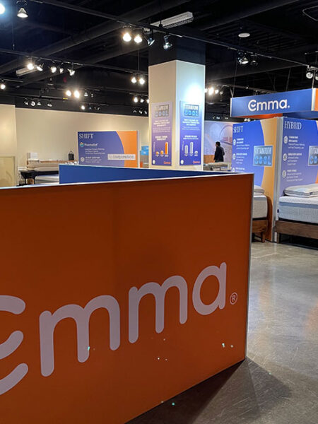 A modern mattress showroom with multiple Emma brand mattresses and prominent orange and blue branding on display.