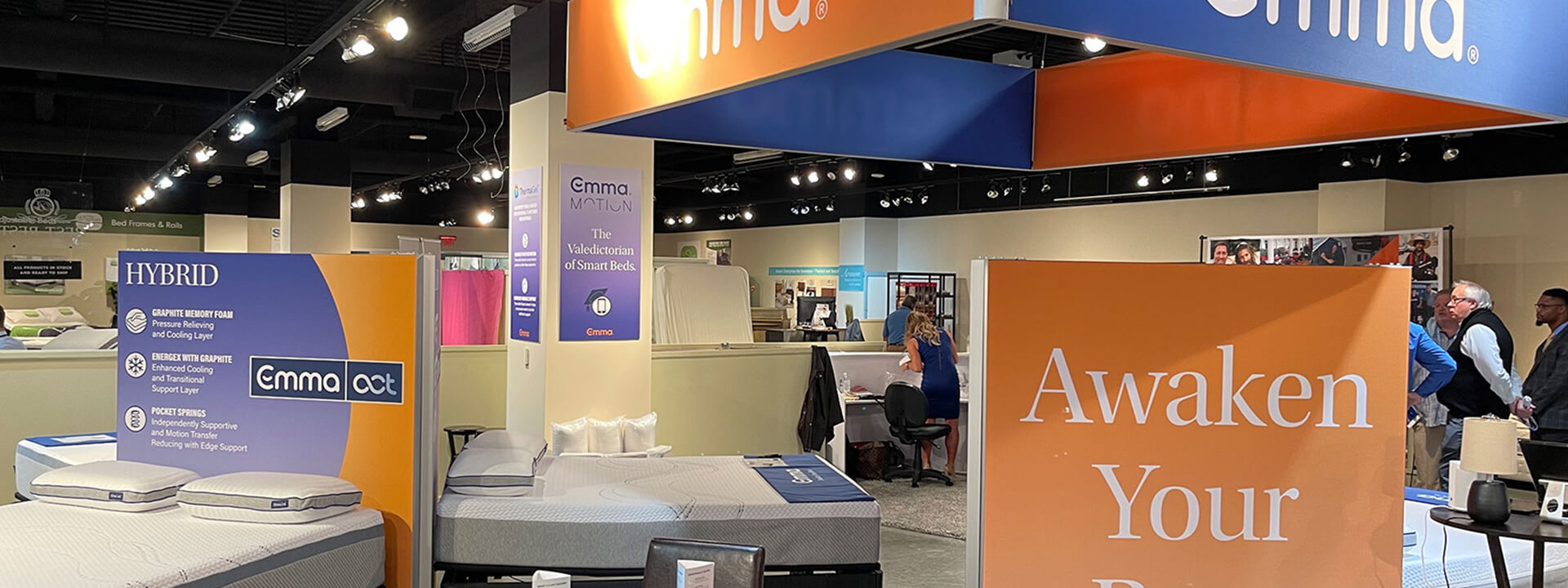 A retail showroom featuring Emma mattresses, informational displays, and people browsing.