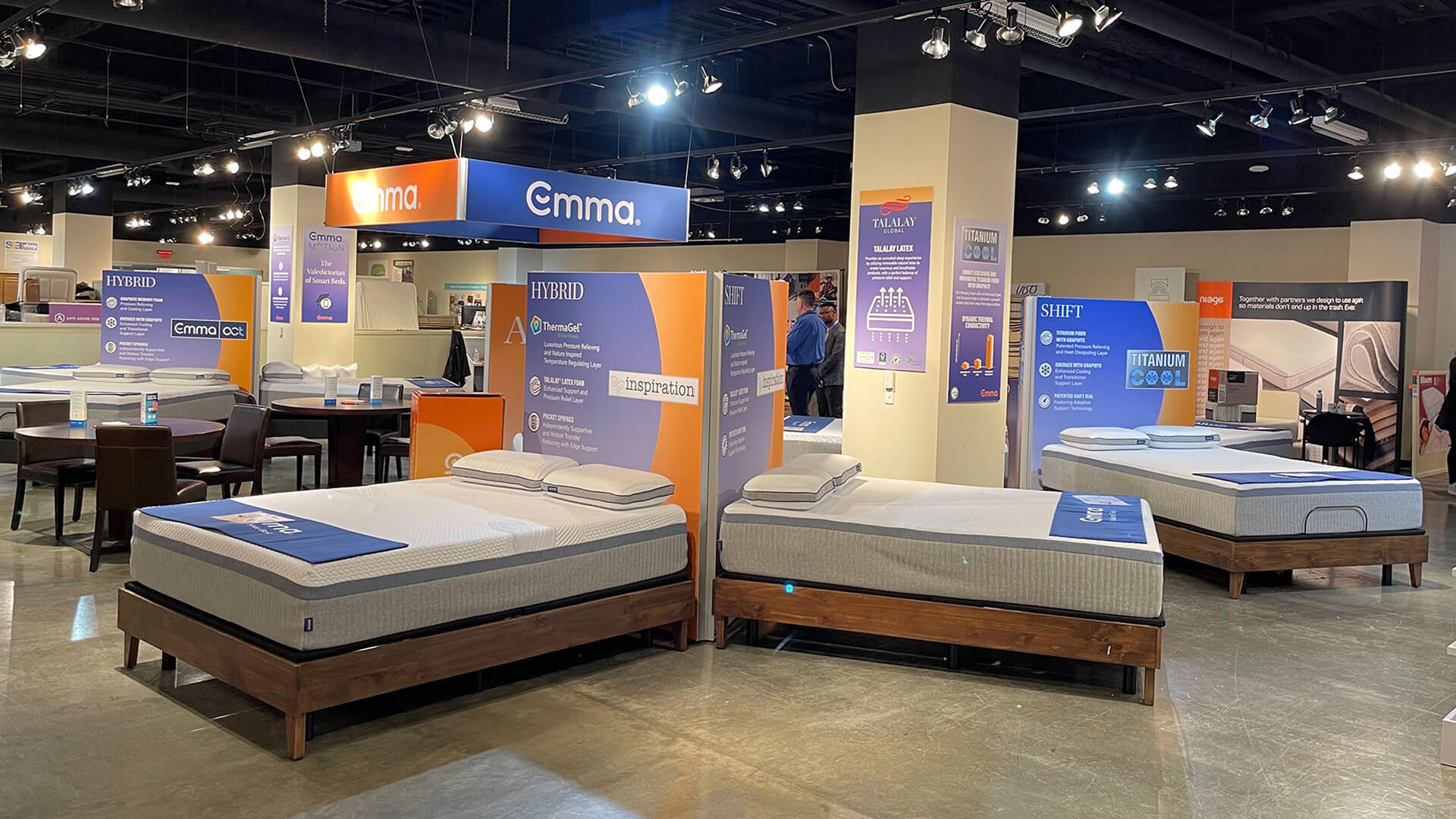 A bright mattress showroom displays several Emma and Hybrid mattresses on wooden bases, surrounded by brand signage.