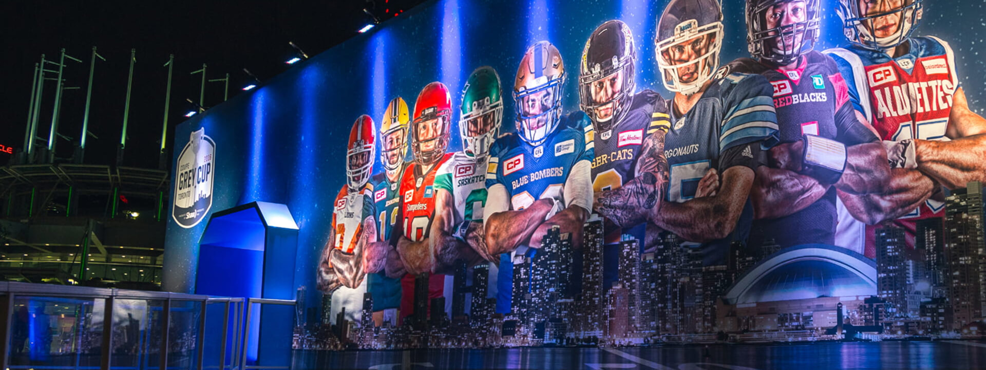 A large outdoor screen at night displays a lineup of football players in various team uniforms above city skylines, with a 2016 Grey Cup Toronto logo on the left.