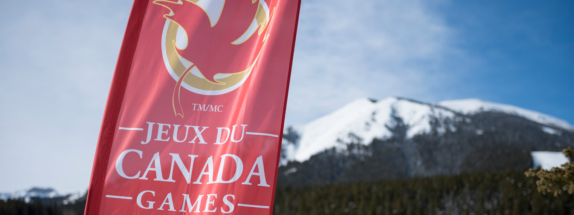 A red banner for the ""Jeux du Canada Games"" with a maple leaf logo stands against a backdrop of a snow-covered mountain and blue sky.