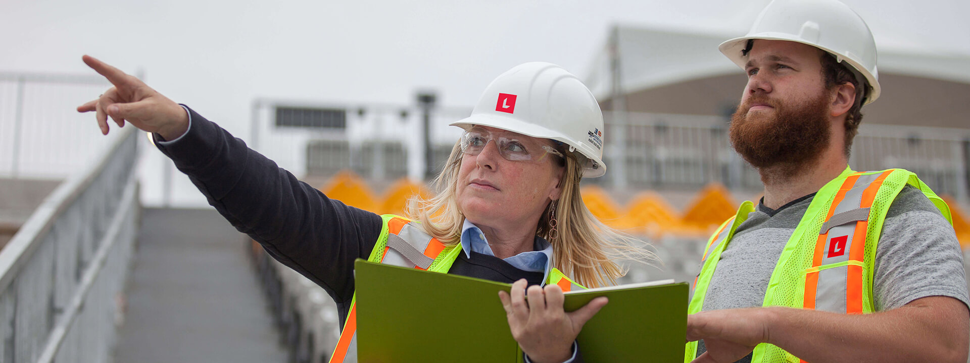 A female and male construction worker, both in hard hats and safety vests, survey a site as the woman points with a folder.