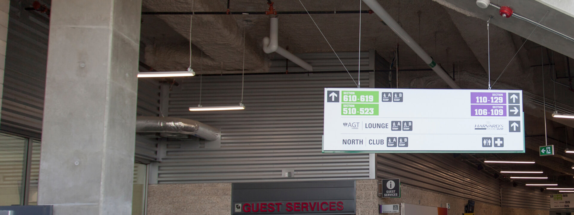 A large white overhead directional sign in an industrial-style interior displays section numbers, lounges, and clubs with arrows.