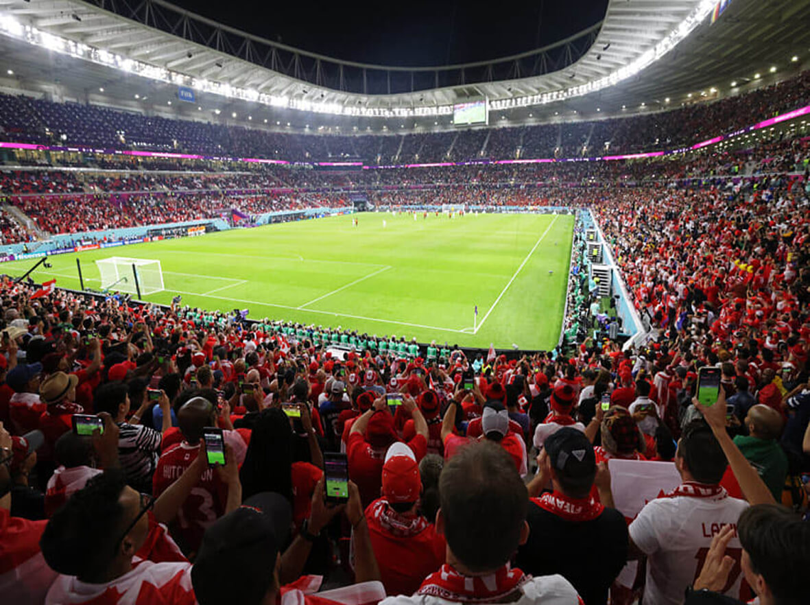A packed stadium at night shows fans in red watching and recording a brightly lit soccer match with their phones.
