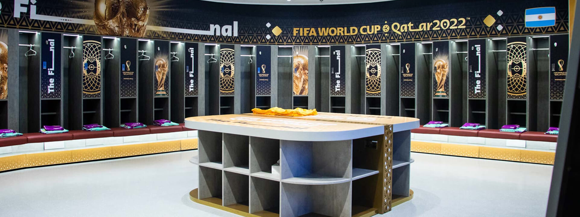 A FIFA World Cup Qatar 2022 final locker room, decorated with the Argentina flag and individual compartments containing jerseys, surrounding a central table.