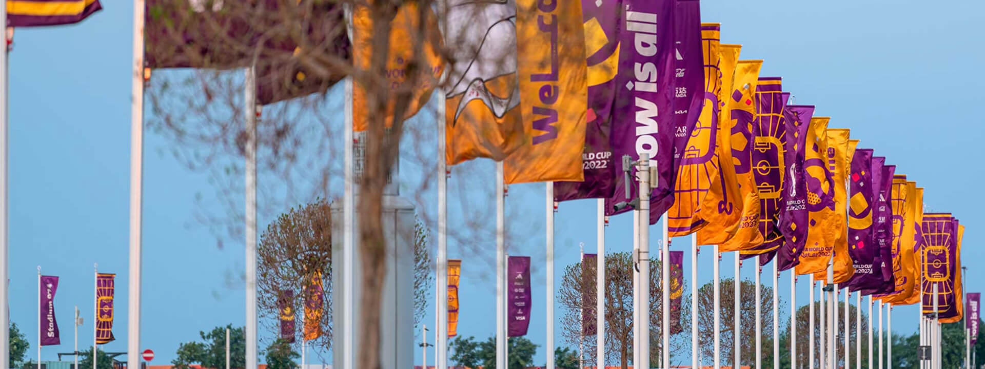 A long row of purple and orange flags for the FIFA World Cup 2022, featuring football stadium graphics and slogans, flies under a clear blue sky.