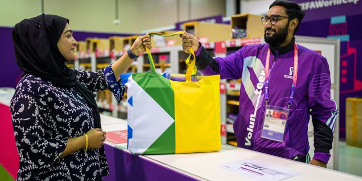 A male volunteer in a purple jacket hands a multi-colored tote bag to a woman wearing a black headscarf.