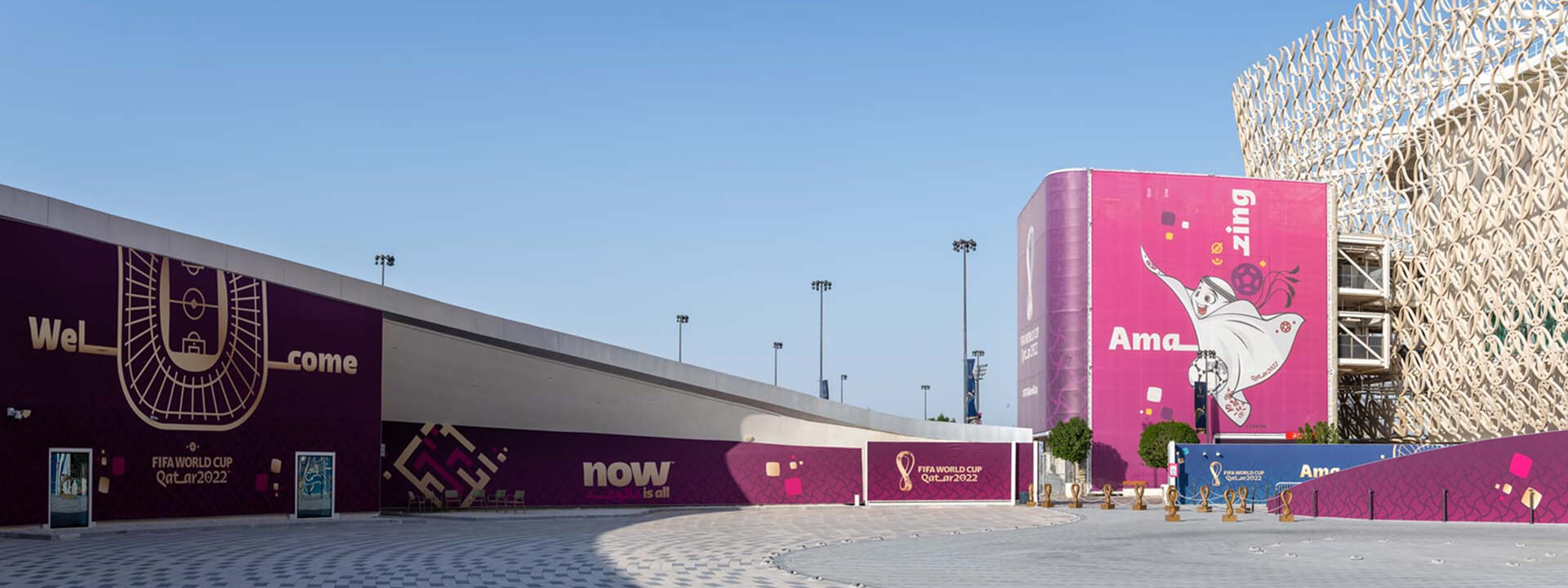 An outdoor plaza is decorated with purple and magenta FIFA World Cup Qatar 2022 signage, featuring ""Welcome"" text and the La'eeb mascot, in front of a modern stadium with a white patterned facade.