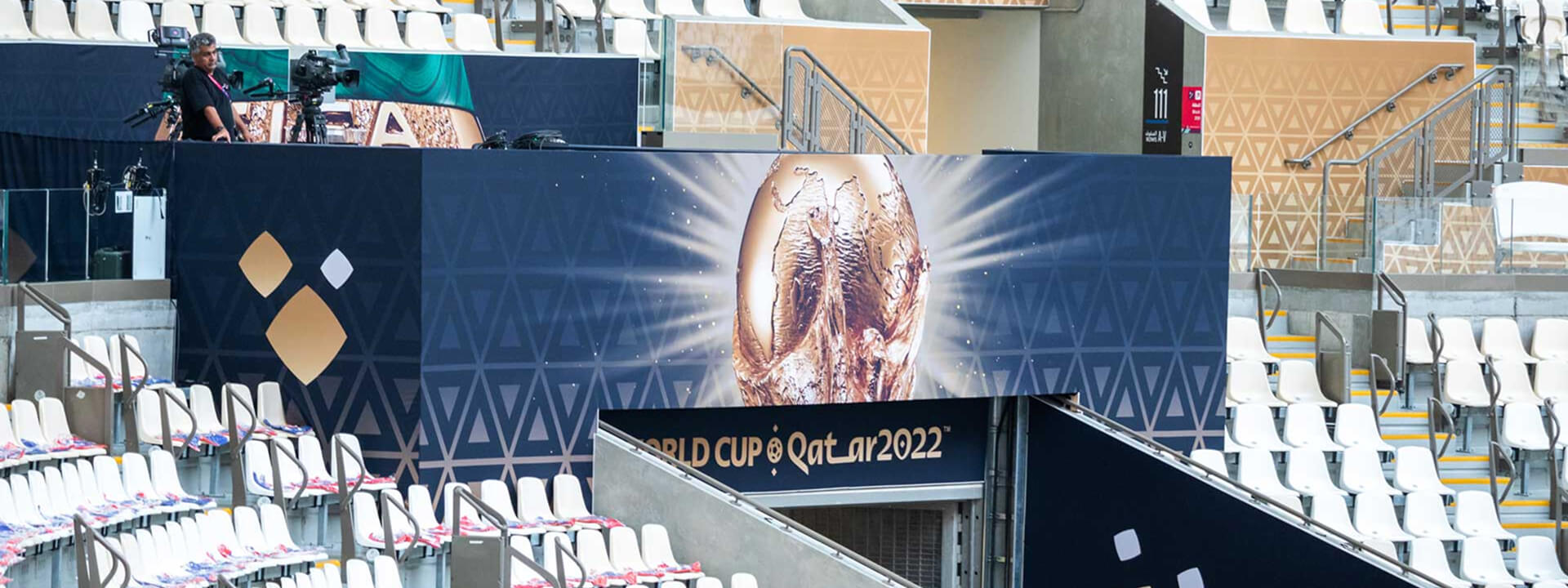 A large banner for the FIFA World Cup Qatar 2022 featuring the golden trophy stands in a stadium with empty seats and a media setup.