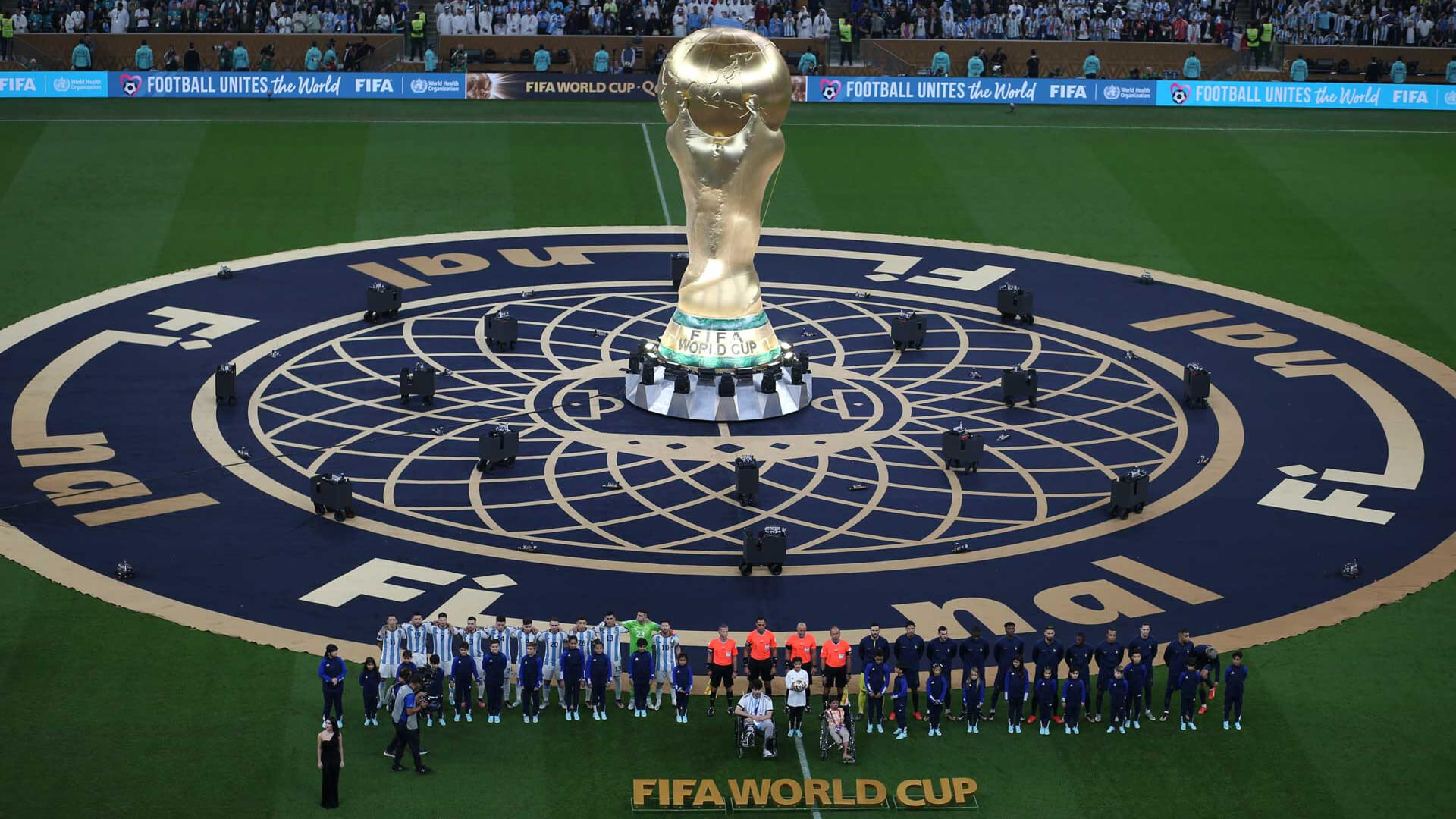 The FIFA World Cup trophy stands on a large circular ""Final"" stage, surrounded by football teams, referees, and children on the stadium pitch.