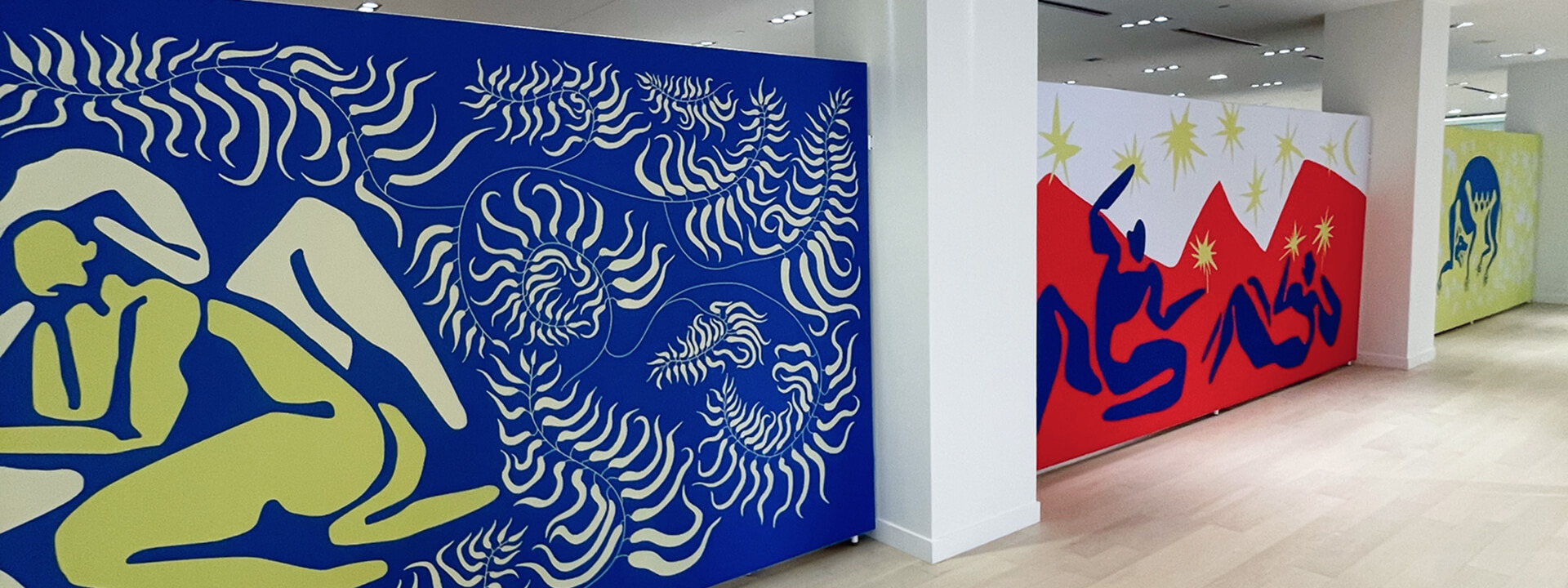 A hallway is lined with large, colorful panels featuring abstract figures and plant shapes reminiscent of Henri Matisse's paper cut-outs.