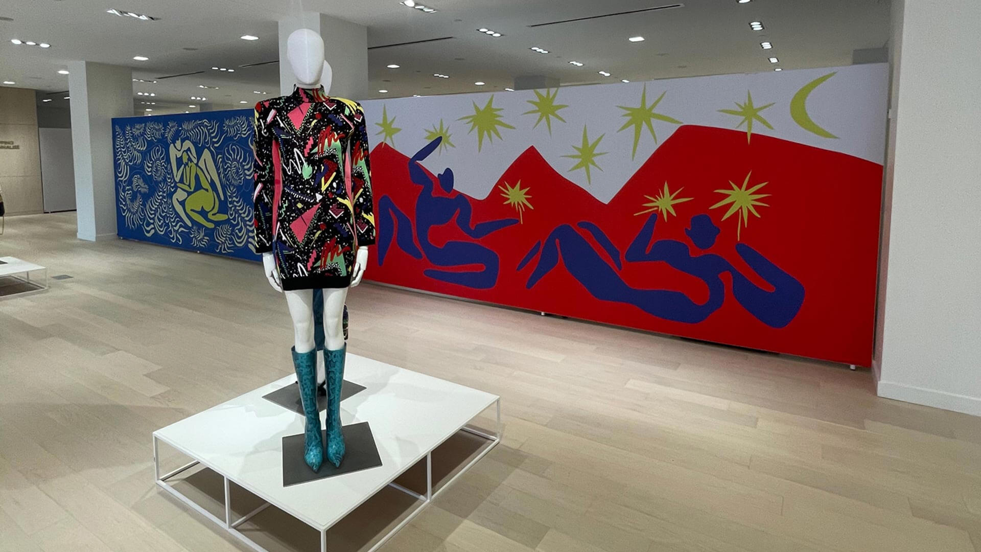 A mannequin in a black patterned dress and teal boots stands before a long wall displaying colorful Matisse-inspired murals.