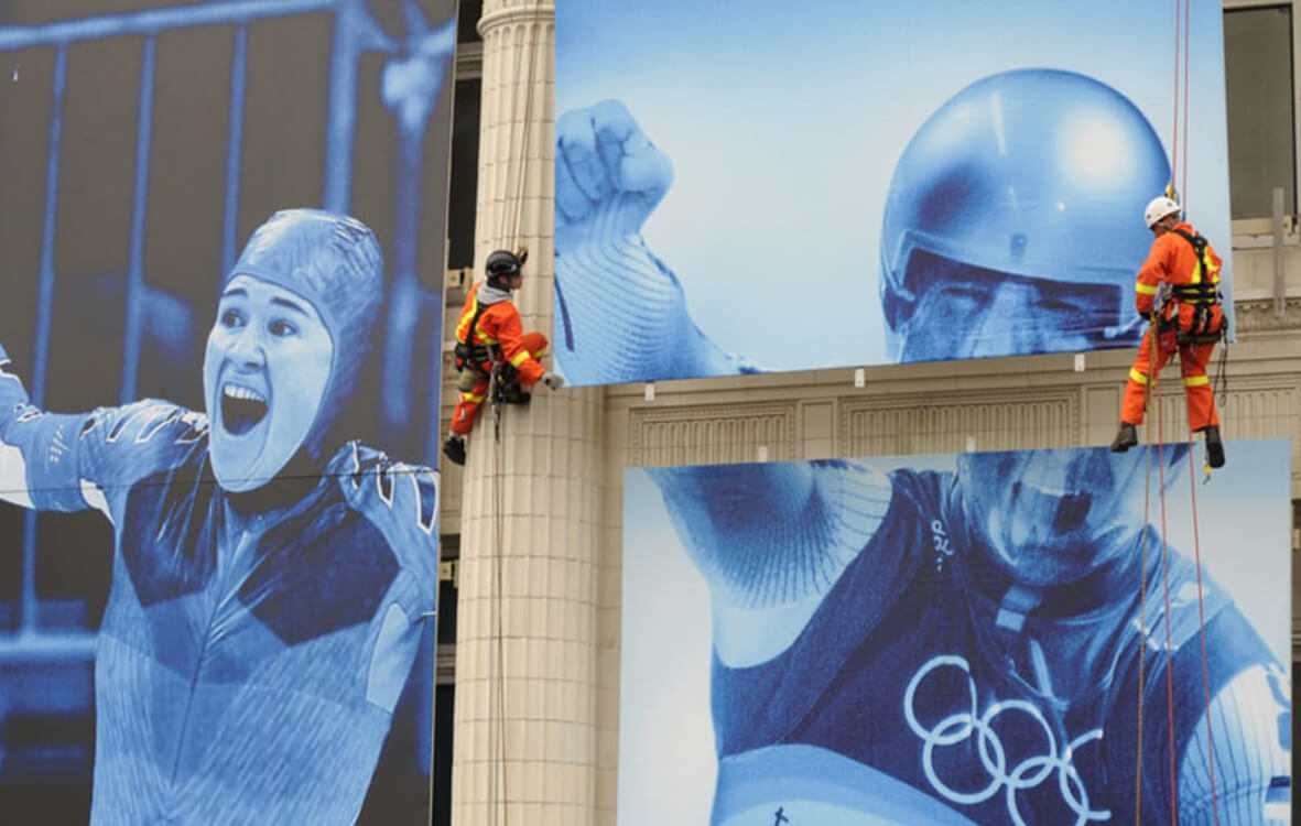 Two workers in orange safety gear rappel down a building displaying giant blue banners of Olympic athletes and symbols.