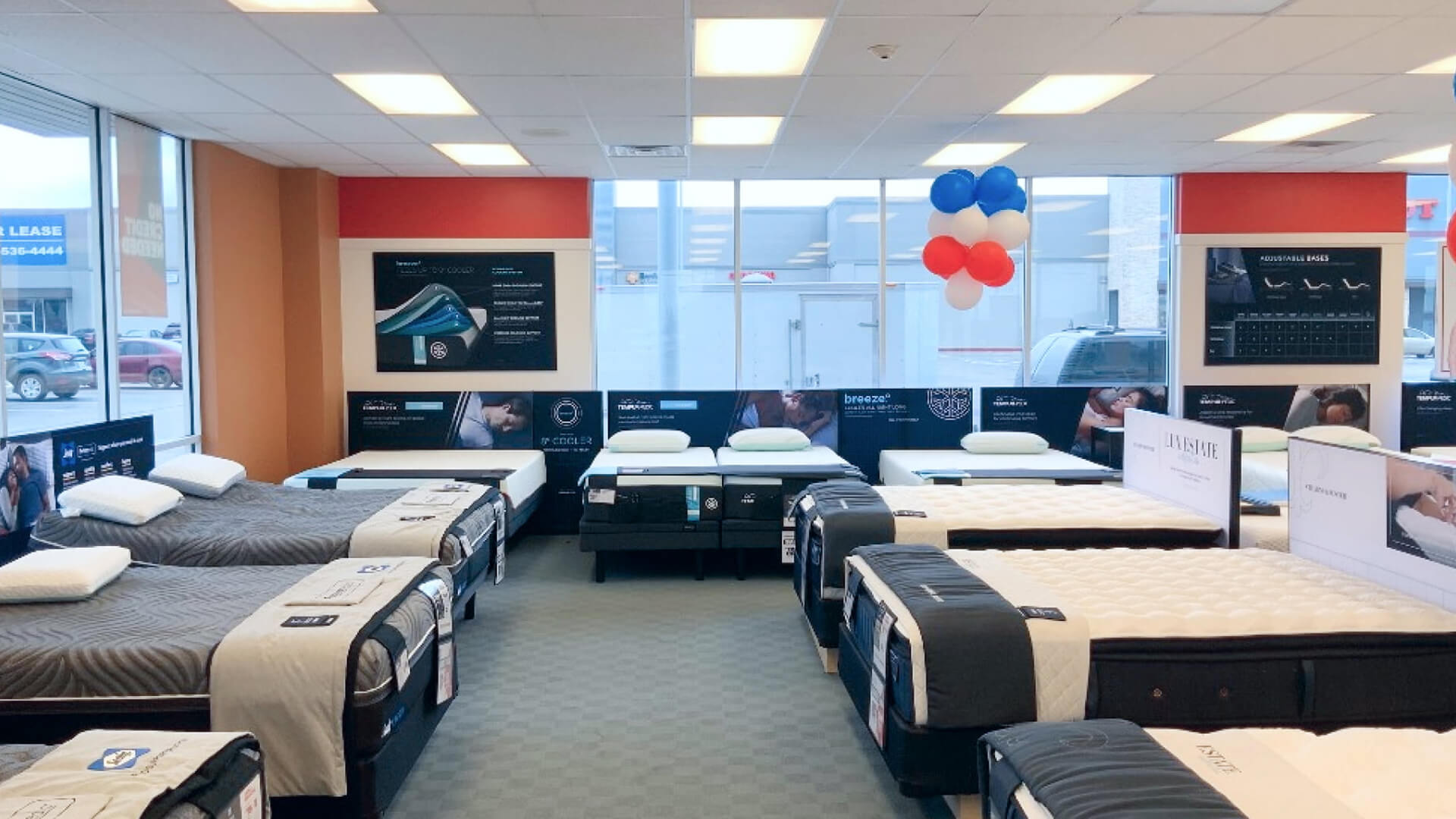 A bright mattress showroom interior features multiple beds with mattresses on display, alongside promotional signage and large windows.