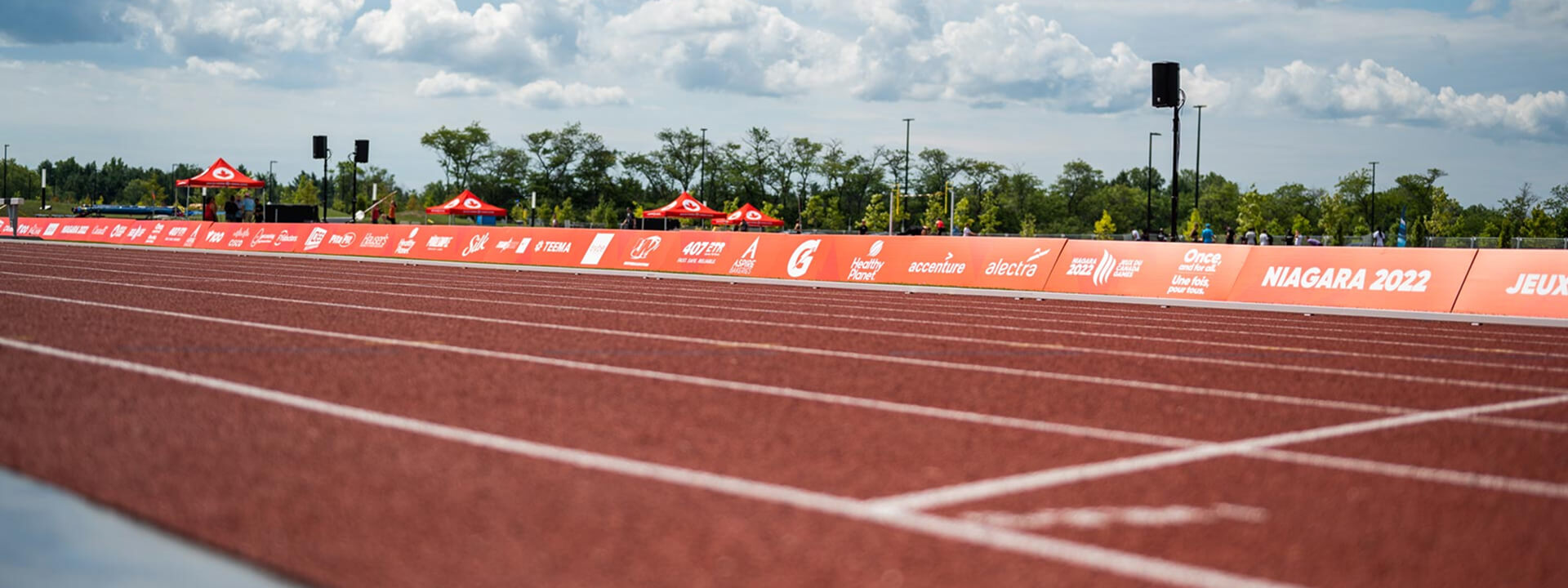 An empty red athletic track runs towards a long orange sponsor banner displaying various logos and ""Niagara 2022"" in an outdoor stadium.