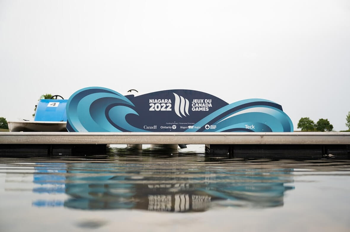 A blue and white sign for the ""Niagara 2022 Jeux du Canada Games"" with a wave design stands on a dock, reflected in the calm water below.