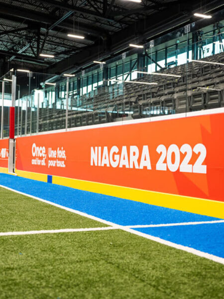 An indoor sports arena features green artificial turf, a blue and yellow track, empty spectator stands, and a large orange banner displaying ""NIAGARA 2022.""
