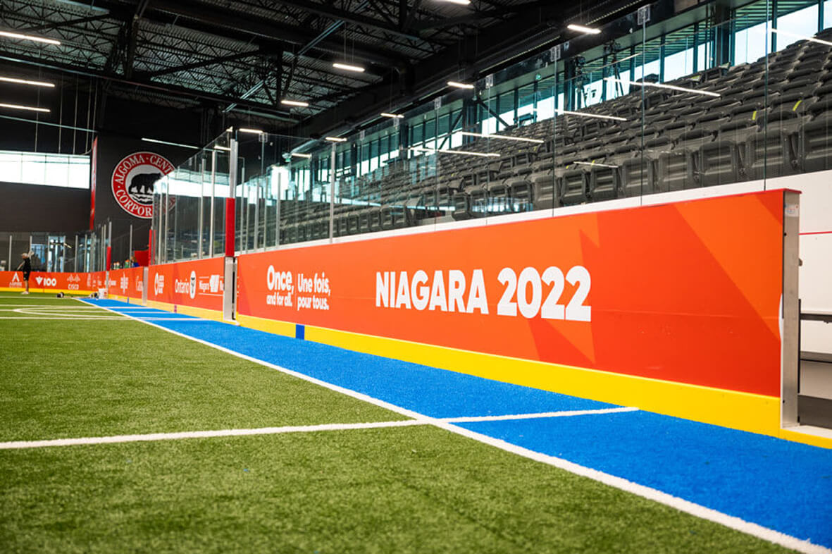 An indoor sports arena features green artificial turf, a blue and yellow track, empty spectator stands, and a large orange banner displaying ""NIAGARA 2022.""