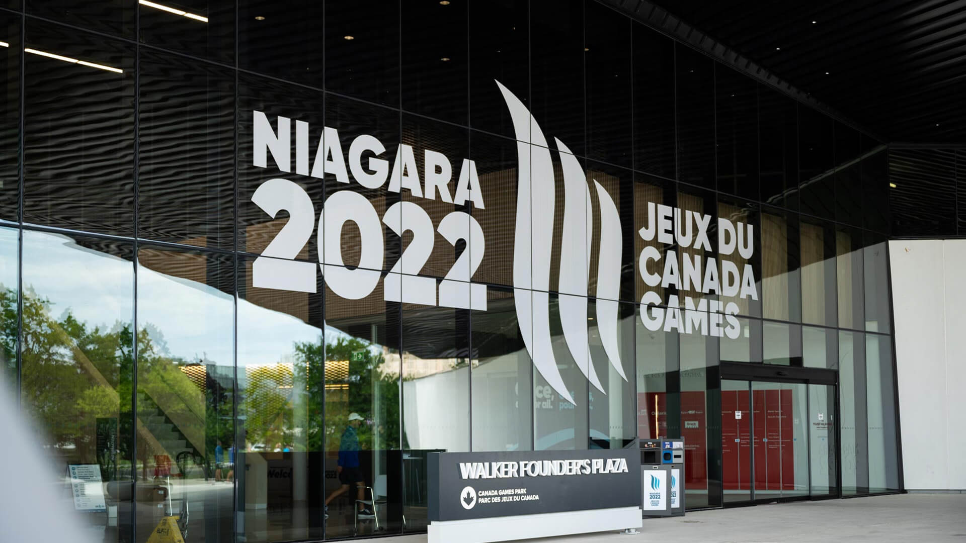 The glass entrance of a modern building displaying ""NIAGARA 2022"" and ""JEUX DU CANADA GAMES"" alongside a ""Walker Founder's Plaza"" sign.