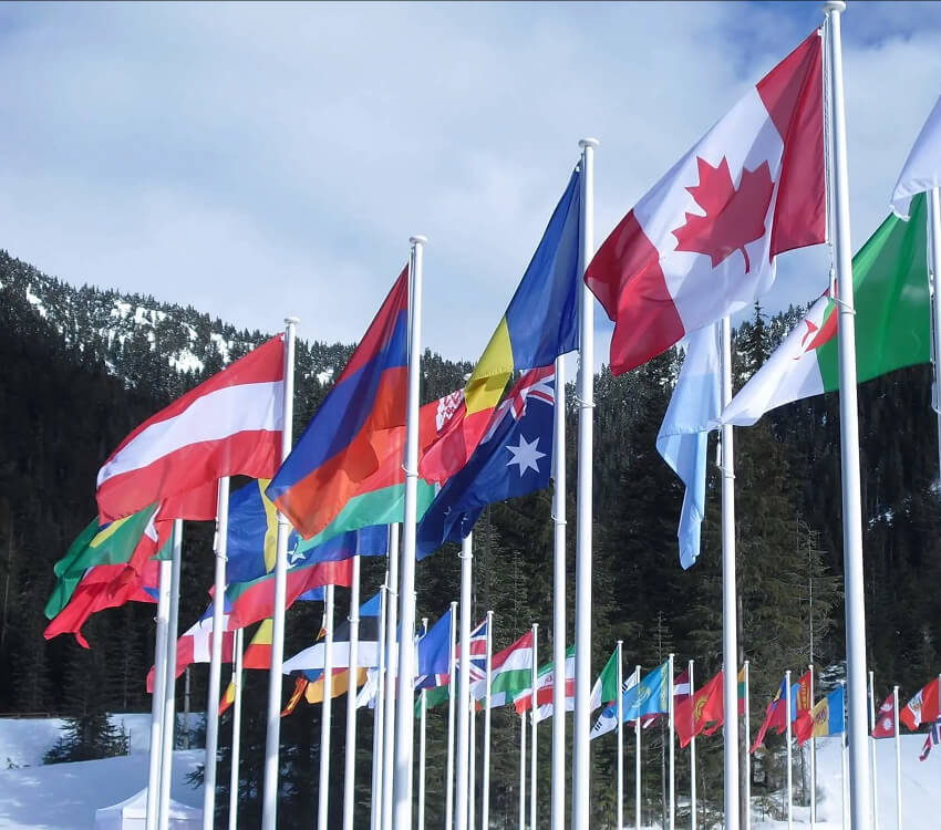 Many international flags fly on poles in a snowy mountain landscape.