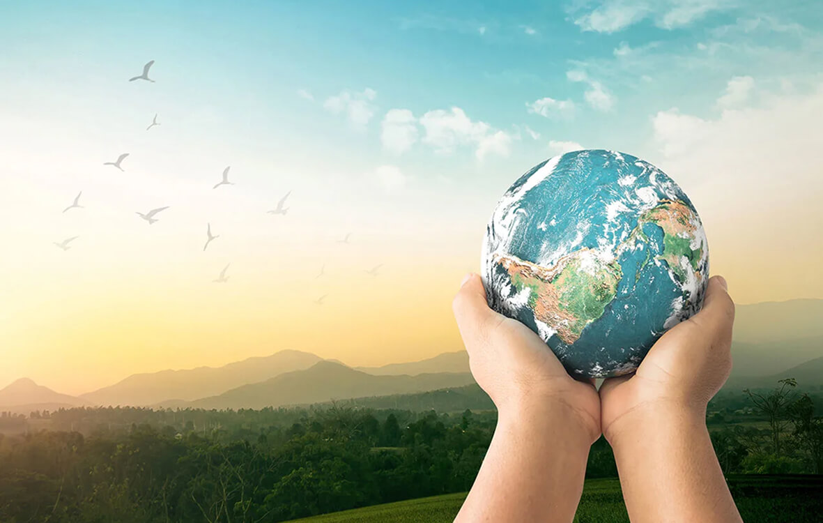 Hands cradle a globe of the Earth, set against a landscape of mountains, forests, and birds flying under a colorful sky.