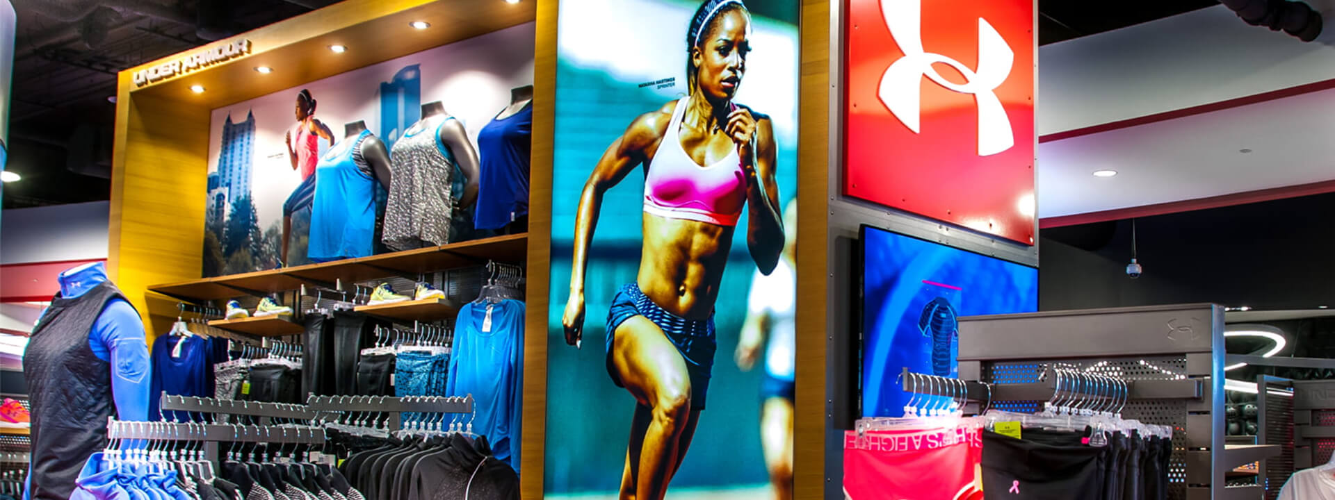 An Under Armour store interior showcasing athletic apparel on mannequins and racks, alongside large displays featuring a female runner and the brand's logo.