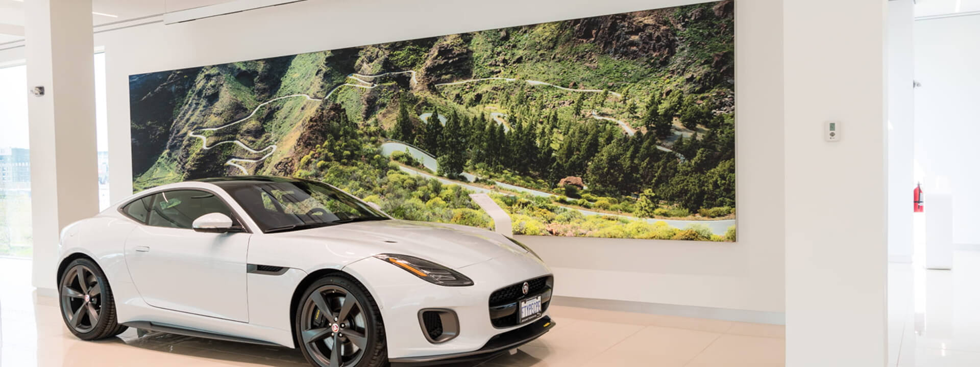 A white Jaguar F-Type sports car is on display in a showroom, with a large mural of a winding mountain road behind it.