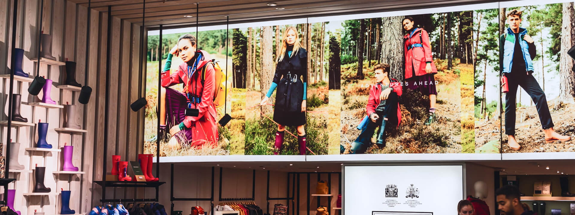 A retail store interior features colorful rain boots displayed on shelves and a large screen showcasing models in outdoor gear within a forest setting.