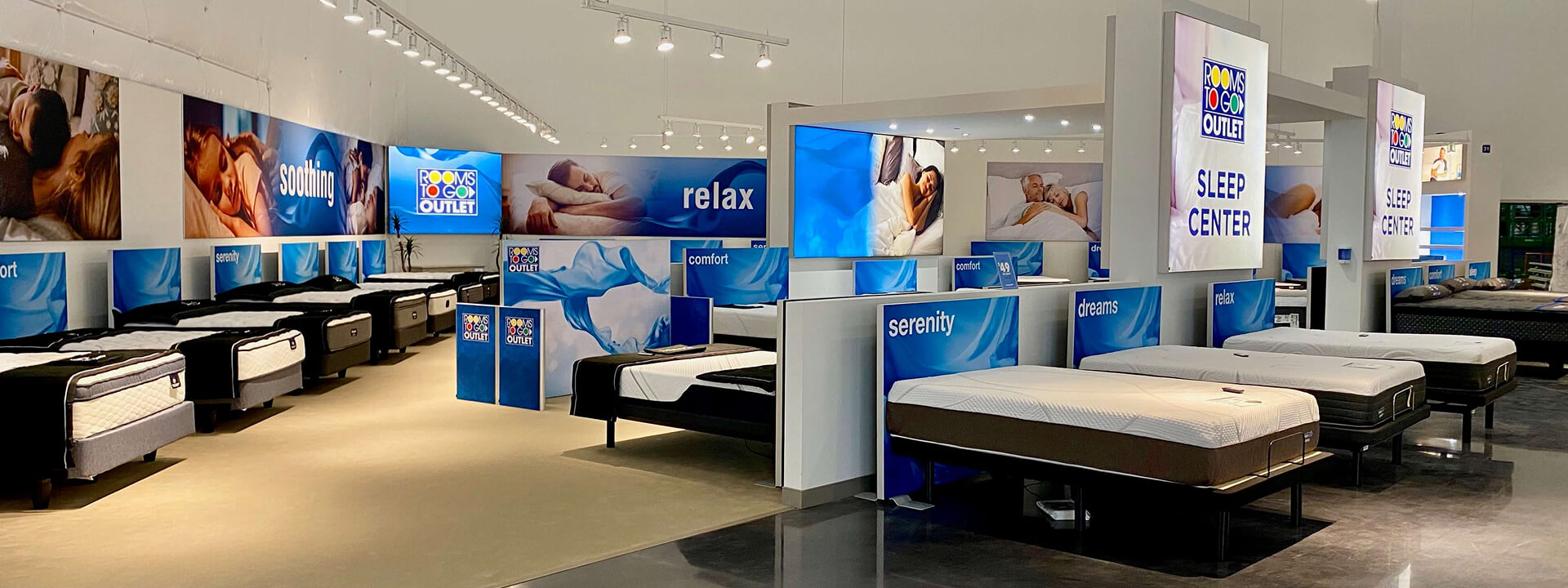 A brightly lit Rooms To Go Outlet Sleep Center showroom features rows of mattresses on display and blue-themed banners with sleeping people and words like ""relax"" and ""soothing.""